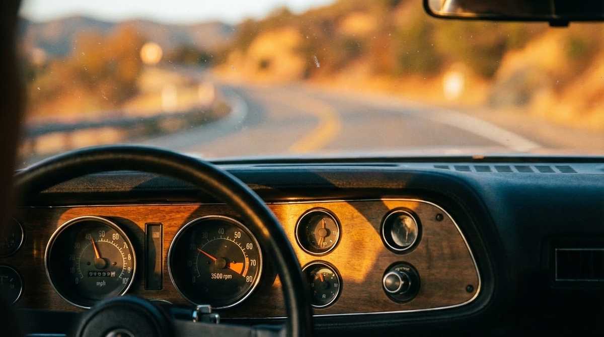 Interior view from the driver’s seat of a car with a vintage dashboard, looking out at a winding road through a sunlit landscape.